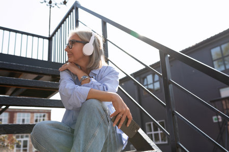 mature woman with gray hair and glasses listens to her favorite playlist in headphones while sitting on the streetの写真素材