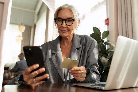 charming mature business woman pays for the order online using a smartphone while sitting in a cafeの写真素材