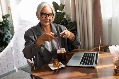 charming stylish mature woman with glasses dressed in a gray jacket sits in a cafe at lunchtime, a laptop lies on the tableの写真素材