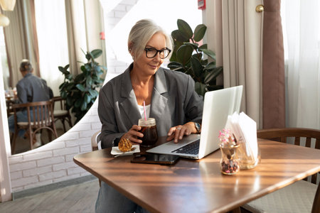 successful well-groomed middle-aged woman with glasses spends time at a laptop while sitting in a cafeの写真素材