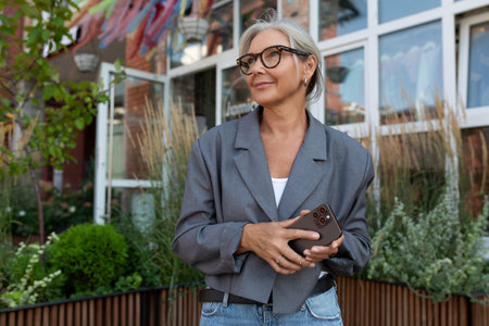 charming slim mature woman with glasses dressed in a gray jacket and jeans holding a smartphone outdoorsの写真素材