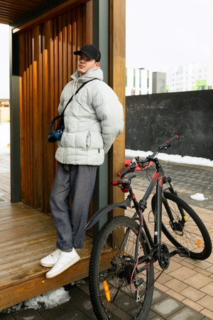 A man in a cap and a warm jacket stands with a rental bicycle