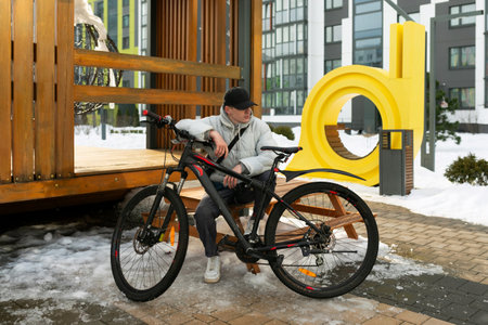 A young Caucasian man with a gray jacket and black cap sits on a bench near a bicycleの写真素材
