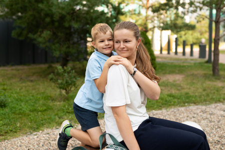 A young boy hugs his mother from behind while they sit together on a bench outsideの写真素材