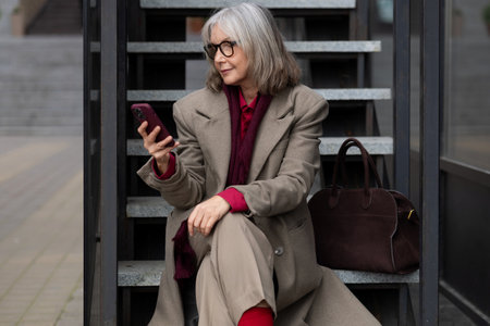 Middle-aged Caucasian businesswoman sitting on stairs, checking her smartphone while dressed in a stylish light-colored coat in an urban settingの写真素材