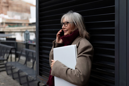 Elderly businesswoman standing outside with a laptop, engaged in a phone conversation in the city during daytimeの写真素材