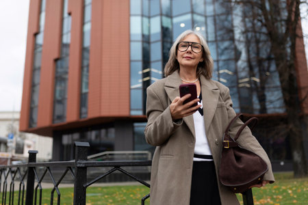 Elderly businesswoman standing with phone outside modern office building on a cool dayの写真素材
