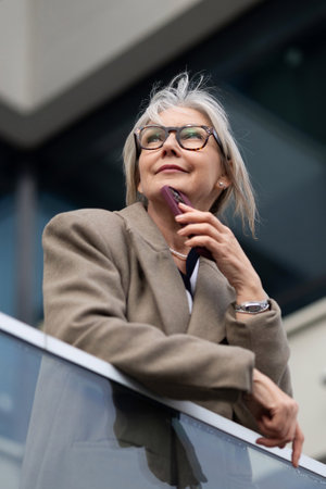 An older woman thoughtfully gazes into the distance while holding her phone on a modern balcony in an urban settingの写真素材