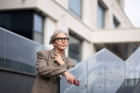 Older woman in stylish glasses stands thoughtfully on a modern staircase in an urban setting during daylightの写真素材