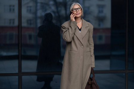 Stylish elderly woman in a long coat standing outdoors talking on the phone near a modern building