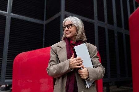 Senior businesswoman stands confidently outside a modern building, holding a laptop, dressed in a...