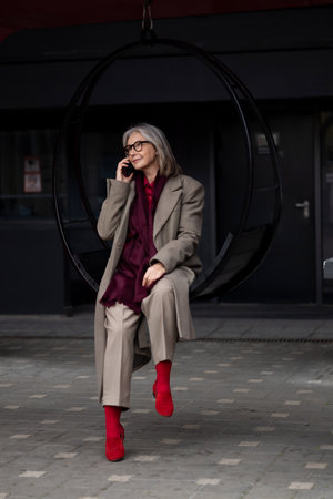 An older woman enjoying a conversation on her phone while seated in a circular swing at a modern outdoor urban settingの写真素材