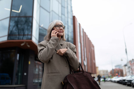 Middle-aged businesswoman with glasses standing outdoors, using smartphone while carrying handbag on a cloudy dayの写真素材