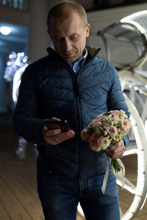 A man walks through the city at night, checking his phone and holding a bouquet of flowersの写真素材