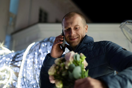 A man walks through the city at night, holding flowers and talking on his phoneの写真素材
