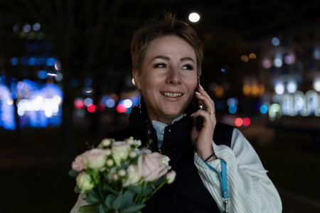 A woman walks through a city at night, talking on the phone and holding a bouquet of flowers