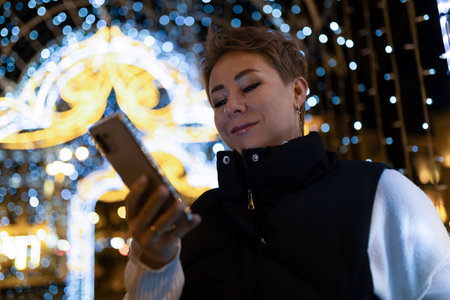 A woman walks through a city at night, looking at her phoneの写真素材