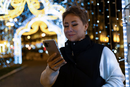 A woman walks through a city at night, looking at her phone, with bright lights illuminating the sceneの写真素材