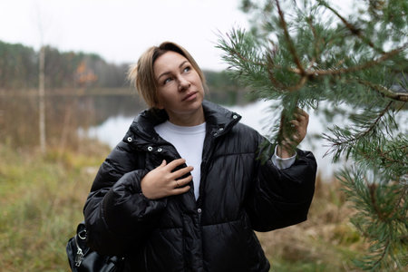 A woman in a black puffer jacket enjoys nature while touching a pine tree near a tranquil lake during an overcast dayの写真素材