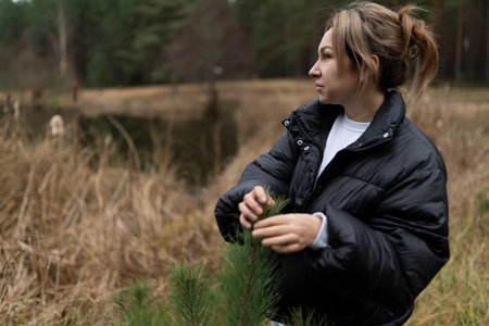 Woman in a black puffer jacket gently holds tree branches while standing near a serene lake in a...