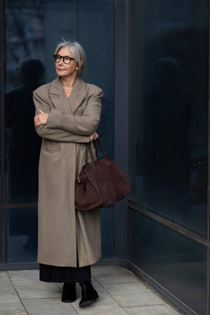 Senior businesswoman confidently standing outside a modern corporate building during daytime, wearing a stylish coat and carrying a handbagの写真素材