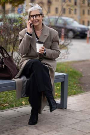 Elderly businesswoman enjoying a coffee while talking on the phone in a park setting during a sunny afternoonの写真素材