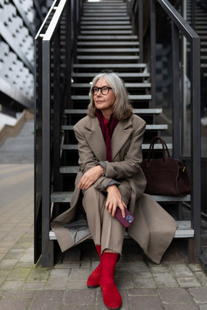 Elderly businesswoman seated on stairs outside an office building during a sunny day, dressed elegantly in a stylish coat and vibrant red accessoriesの写真素材
