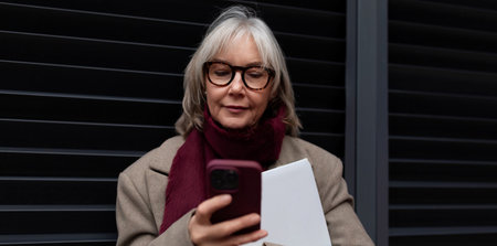Elderly businesswoman standing outside her office in winter attire while checking her smartphone...