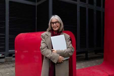 Elderly businesswoman confidently standing outside her office, holding a folder, surrounded by...