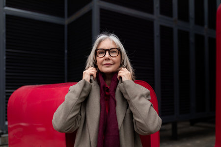 An older woman adjusts her scarf against a red backdrop in an urban setting during a cool afternoon