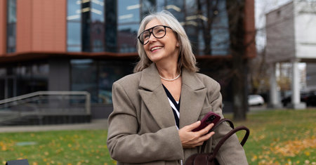 An older woman smiles while walking in the park near a modern building on a sunny day during autumnの写真素材