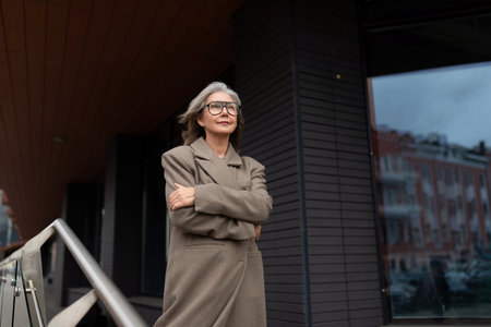 Older woman in a stylish coat stands confidently outside a modern building on a sunny day, enjoying the moment and the viewの写真素材