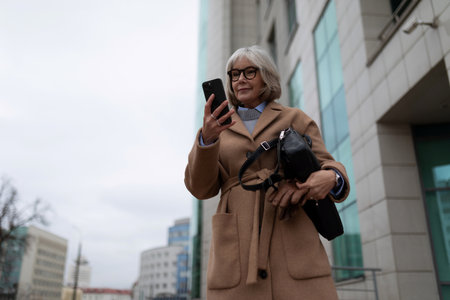 Elderly woman dressed in a stylish coat checks her smartphone while standing outside a modern building on an overcast dayの写真素材