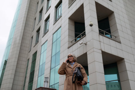 Woman talking on the phone while standing outside a modern building in a busy urban area during cloudy weatherの写真素材