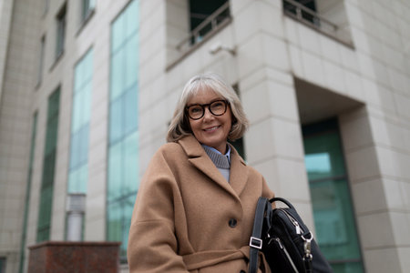 Smiling senior woman with glasses poses outside a modern building in autumn, showcasing her stylish...