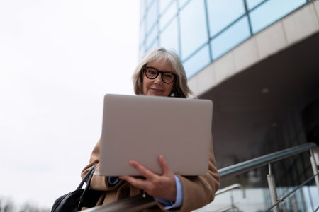 Senior woman using a laptop outdoors in front of a modern building during the day, showcasing work and connection in urban lifeの写真素材