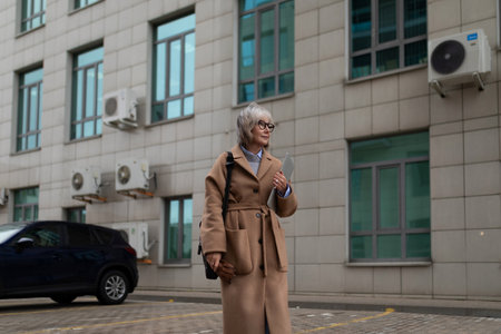 Stylish older woman walks outside a modern building on a cool day in the city, showcasing a fashionable brown coat and confident demeanorの写真素材