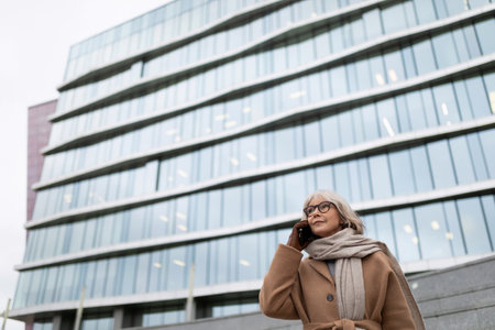 Elderly woman in a stylish coat talking on the phone outside a modern glass building on a cool dayの写真素材