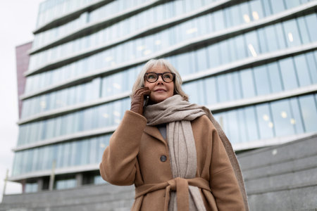 A woman in a stylish coat talks on her phone in front of a modern office building on a chilly dayの写真素材