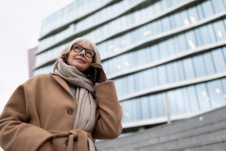 An elderly woman talks on her smartphone while standing outside a modern building on a chilly dayの写真素材