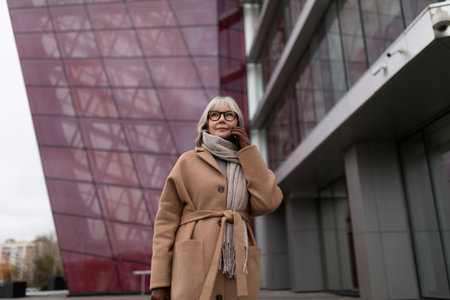 Woman in stylish coat and scarf standing near modern architecture on a cloudy day in an urban setting during autumnの写真素材
