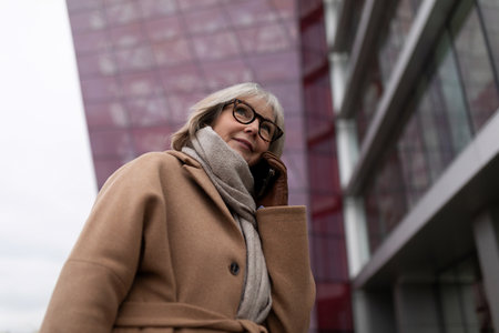 An older woman in a tan coat talks on her smartphone near a modern building in a city during a cloudy dayの写真素材