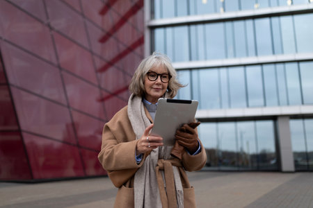 Senior woman using digital tablet outdoors in modern urban setting with glass buildings during overcast dayの写真素材