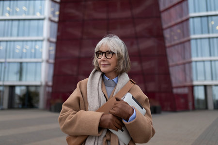 Mature woman in a stylish coat holding a laptop outside a modern building during daytime in an urban settingの写真素材