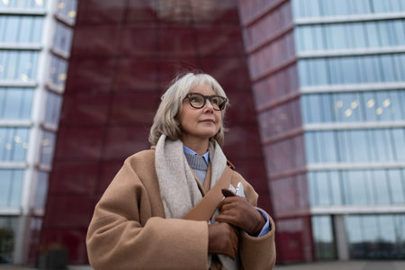 An elderly woman with glasses poses confidently in front of a modern building with glass and red walls during the dayの写真素材