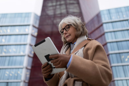 Senior woman in stylish coat reading tablet outside modern glass building during late afternoonの写真素材