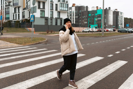 Woman crossing the street while talking on her phone in a residential area with modern buildingsの写真素材