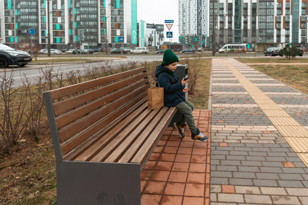 Young boy sitting on a park bench with a smartphone in a modern urban area during a cloudy dayの写真素材