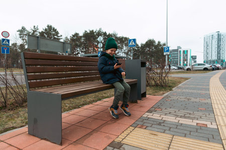 Young boy sitting on a bench in an urban area while using a smartphone on a chilly dayの写真素材