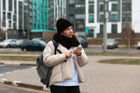 Young woman waits on the street while checking her phone in a modern urban area during a chilly dayの写真素材